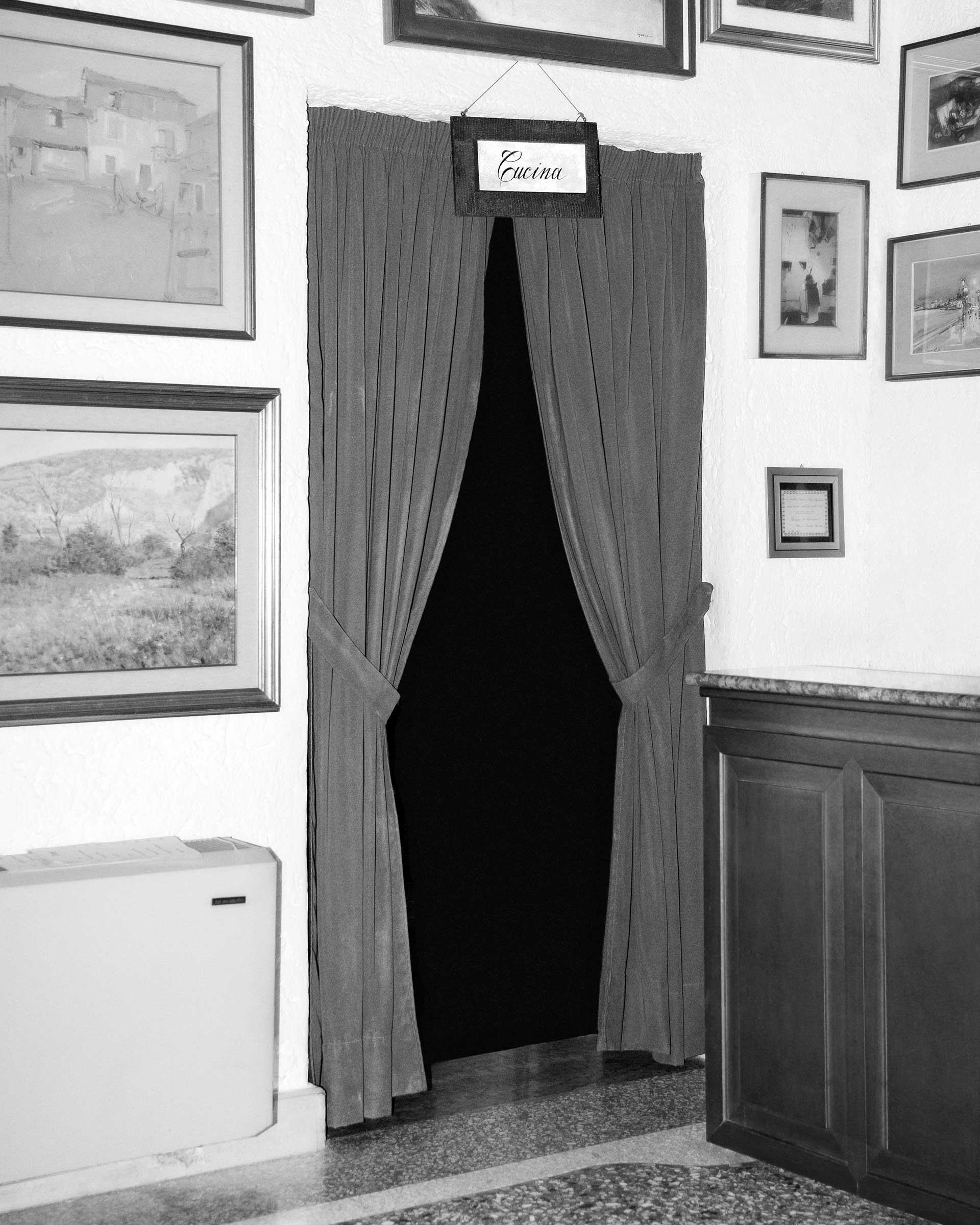 Corner of a room with paintings on the walls and entryway to a kitchen with curtains